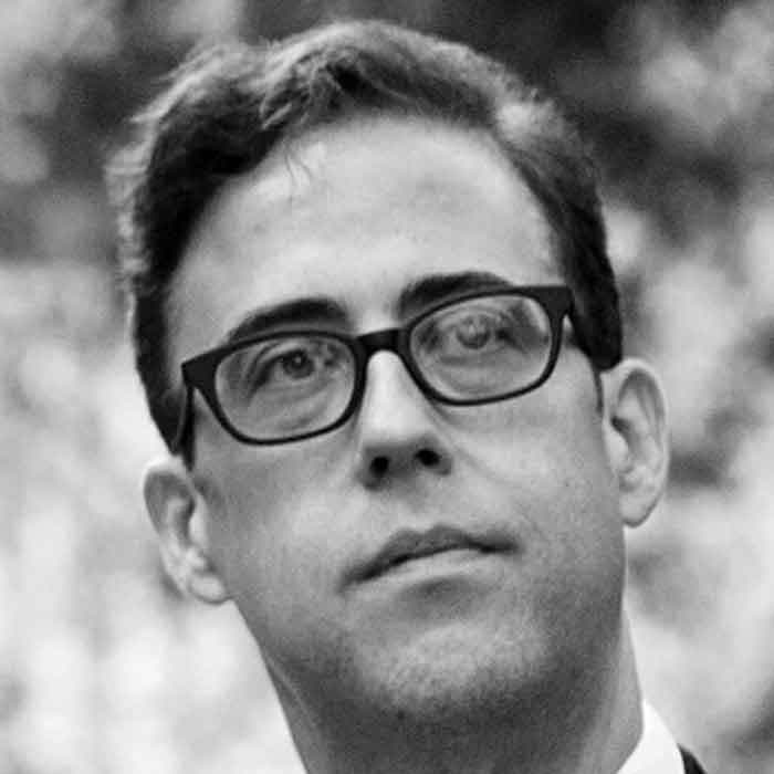 A black-and-white photo of a person with short, dark hair and glasses. The person is looking slightly to the left. The background is blurred, highlighting the subject's face.