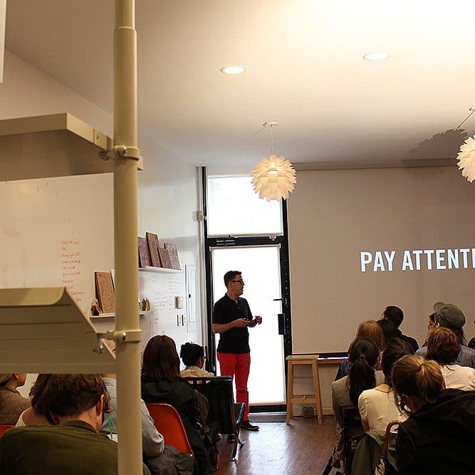 A person giving a presentation stands in front of a seated audience. The projected slide behind reads "PAY ATTENTION." The room has white walls, a large window in the back, and hanging light fixtures. A whiteboard with notes is visible on the left side.