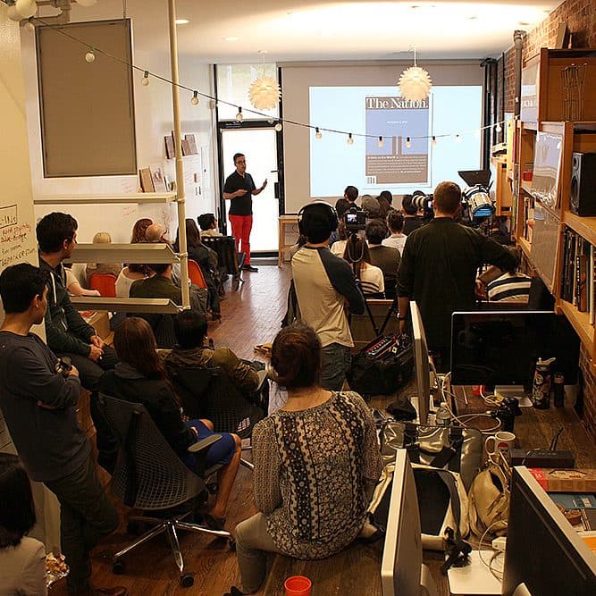 A group of people sit and stand in a room, attentively listening to a speaker who is presenting information on a projected screen. The room has bookshelves, a whiteboard, and various pieces of office equipment, creating an informal meeting or workshop atmosphere.