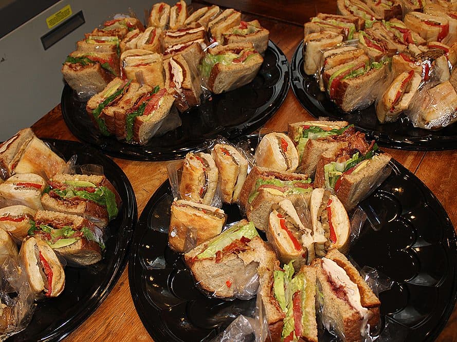 Several black plastic trays are filled with various sandwiches, each cut in half and wrapped in clear plastic. The sandwiches contain ingredients like lettuce, tomato, meat, and cheese, displayed on a wooden surface.