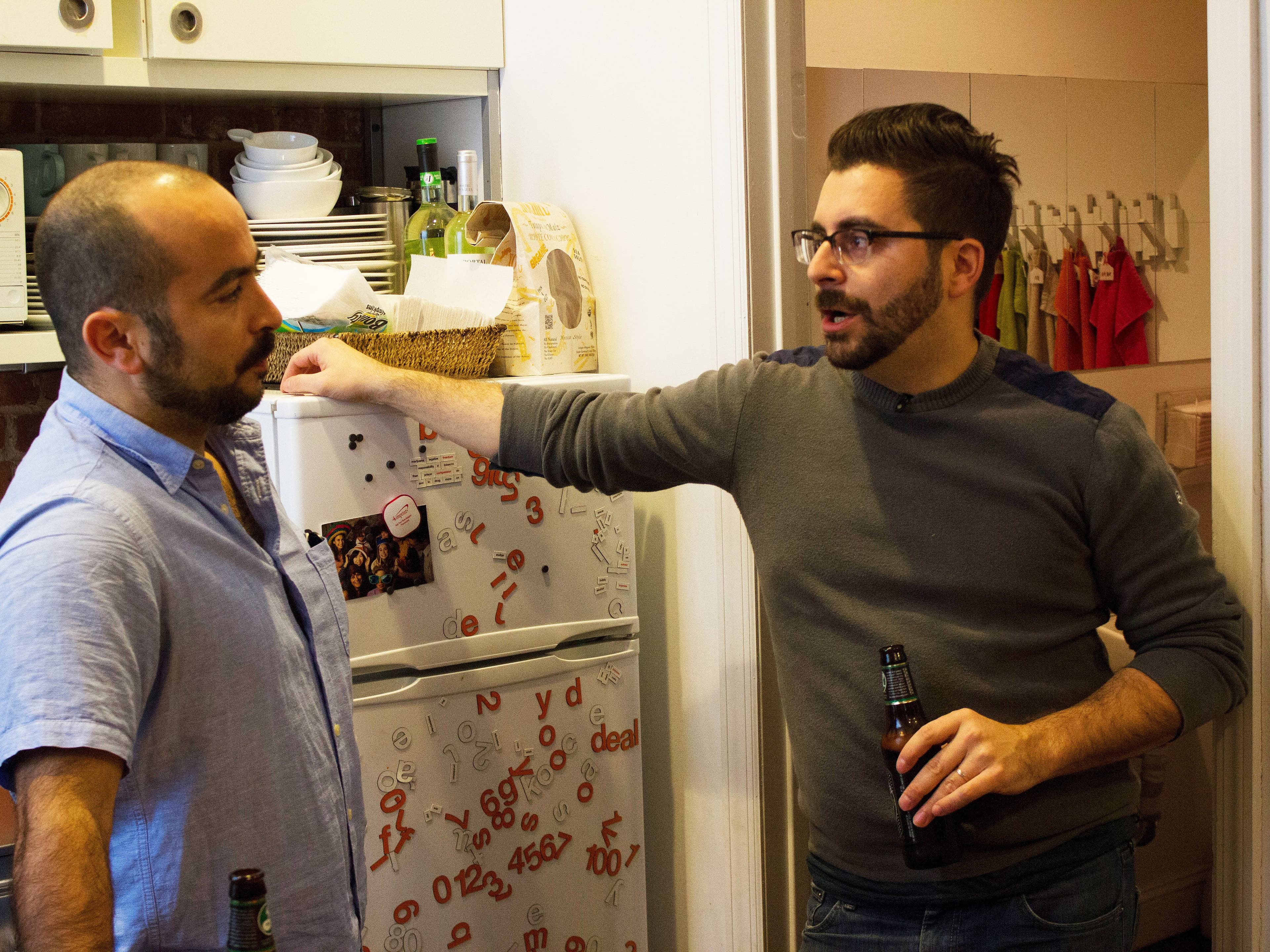 Two men are standing in a kitchen, having a conversation. One man in a blue shirt is leaning against a white refrigerator, while the other man in a gray sweater is gesturing with a bottle in hand. The kitchen is cluttered with various items and papers.