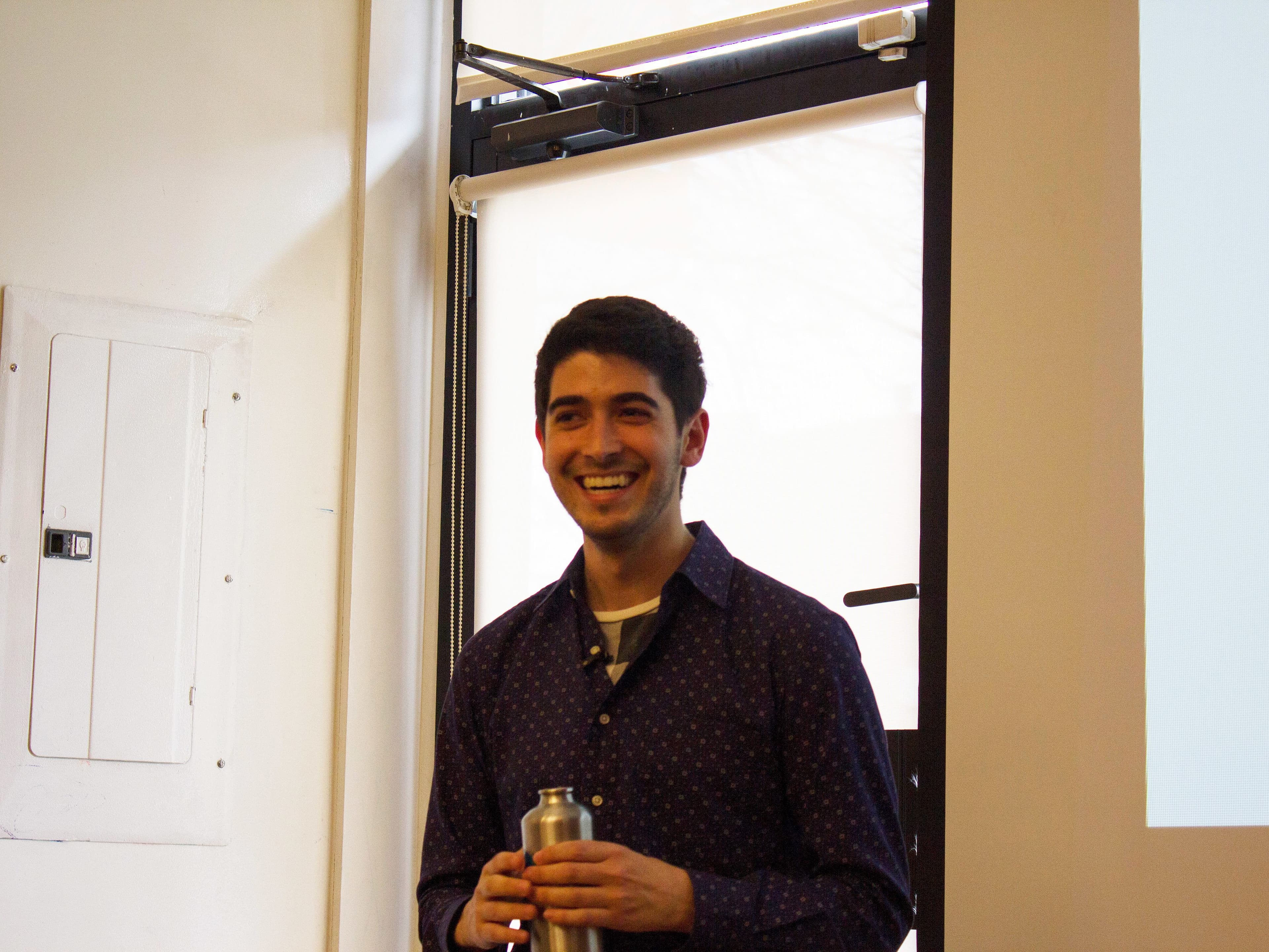 A person with short dark hair stands indoors, smiling and holding a silver tumbler. They are wearing a patterned dark blue shirt. The background features a window with blinds and part of a white wall with an electrical panel visible.