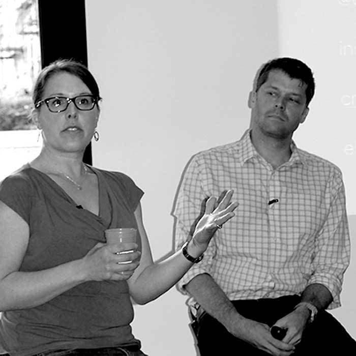 A woman wearing glasses and holding a cup speaks while gesturing with her hand. A man in a plaid shirt sits beside her, looking ahead. They appear to be participating in a discussion or presentation. The setting is indoors with a window in the background.