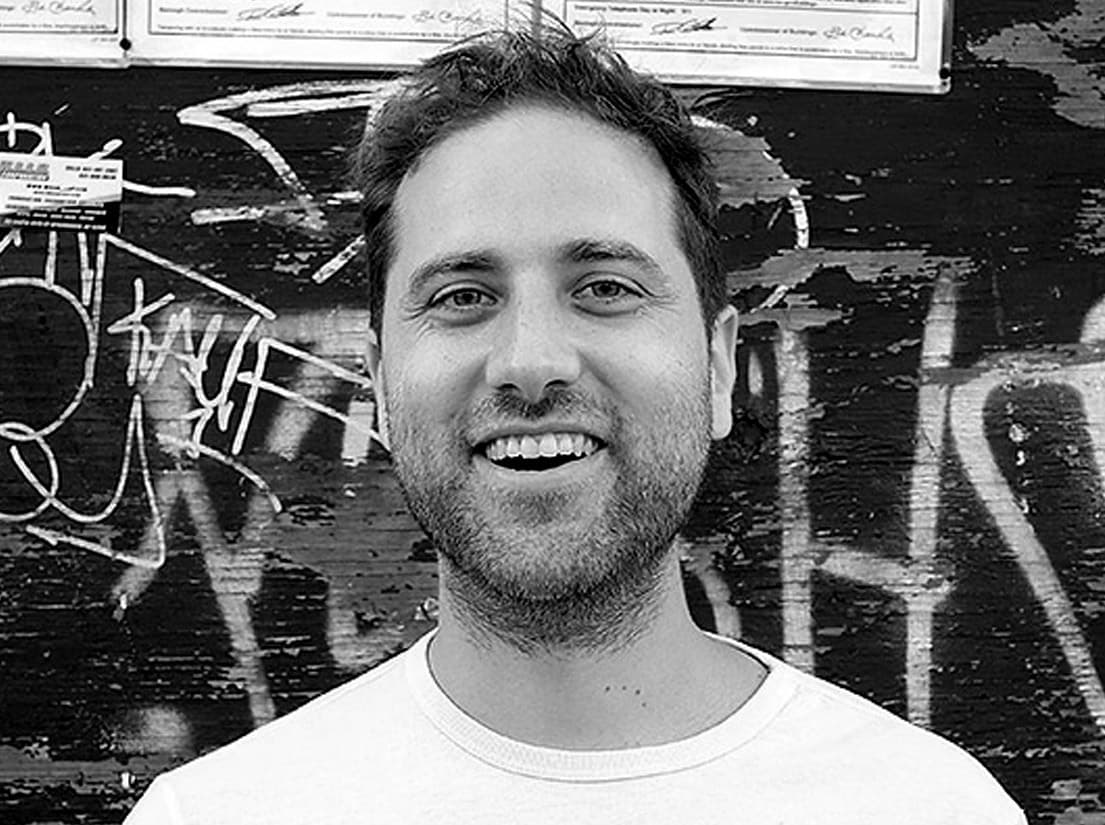 A man with short hair and a beard is smiling at the camera. He is wearing a light-colored shirt and standing in front of a wall covered in graffiti and flyers. The image is in black and white.