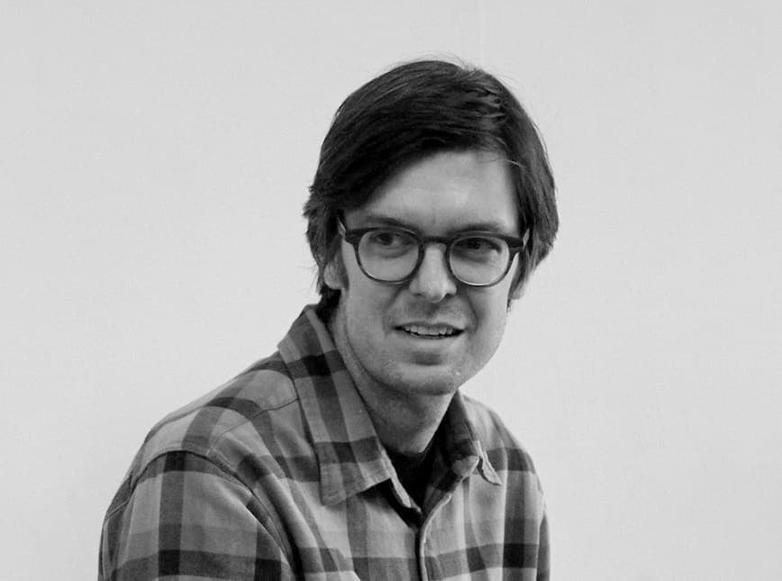 A person with short dark hair and glasses is looking slightly to the side. They are wearing a checkered shirt and appear to be in a neutral setting, against a plain background.
