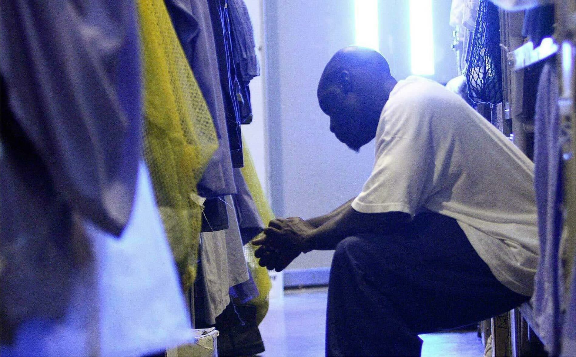 A man sits on a bench in a locker room, leaning forward with his elbows resting on his knees and hands clasped together. He appears deep in thought, surrounded by hanging items of clothing. The lighting is dim with a bluish tint.