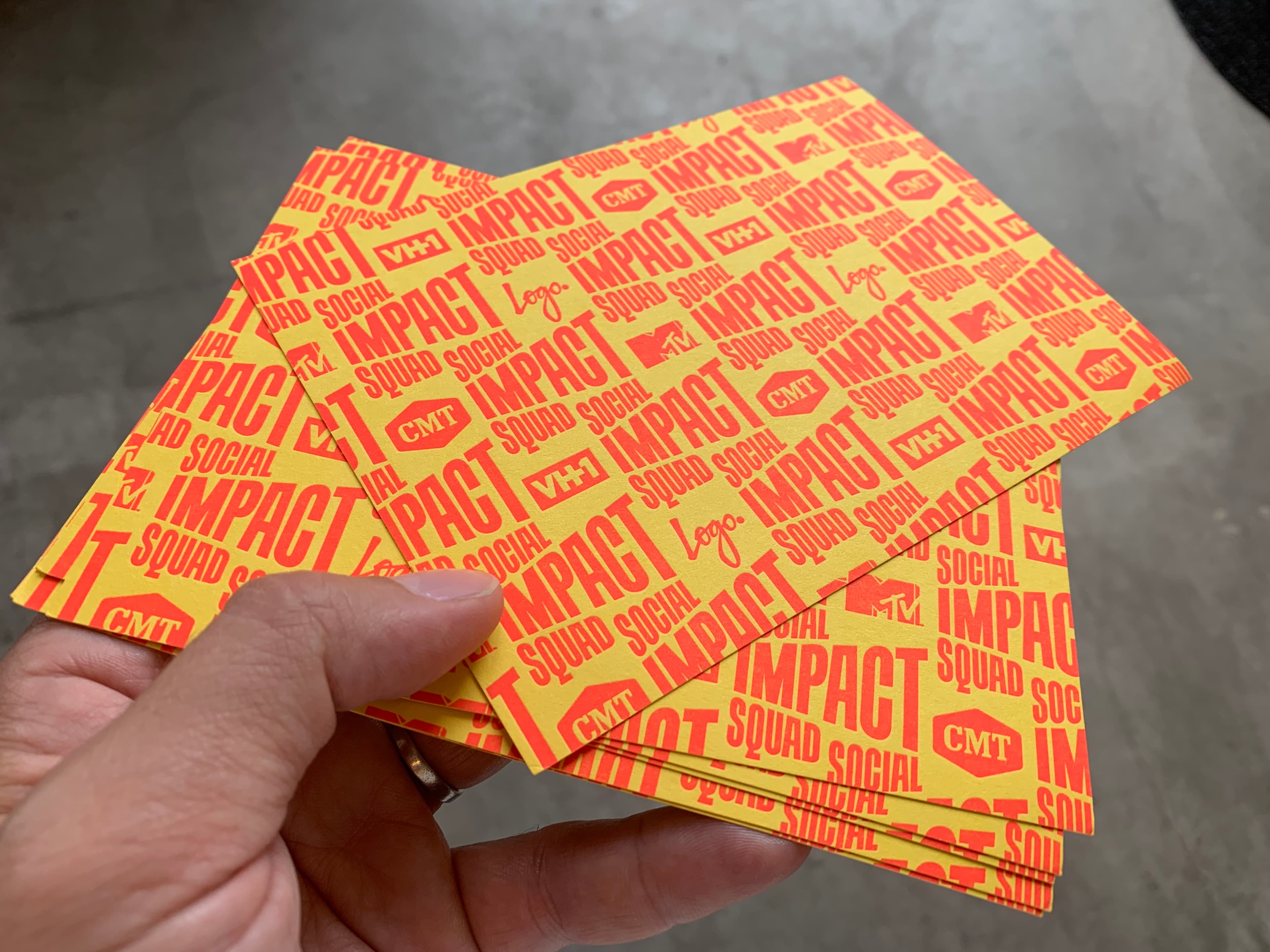 A hand holds several orange and yellow cards filled with words and logos, including "IMPACT," "SOCIAL SQUAD," and logo images of VH1, MTV, and CMT against an urban background.