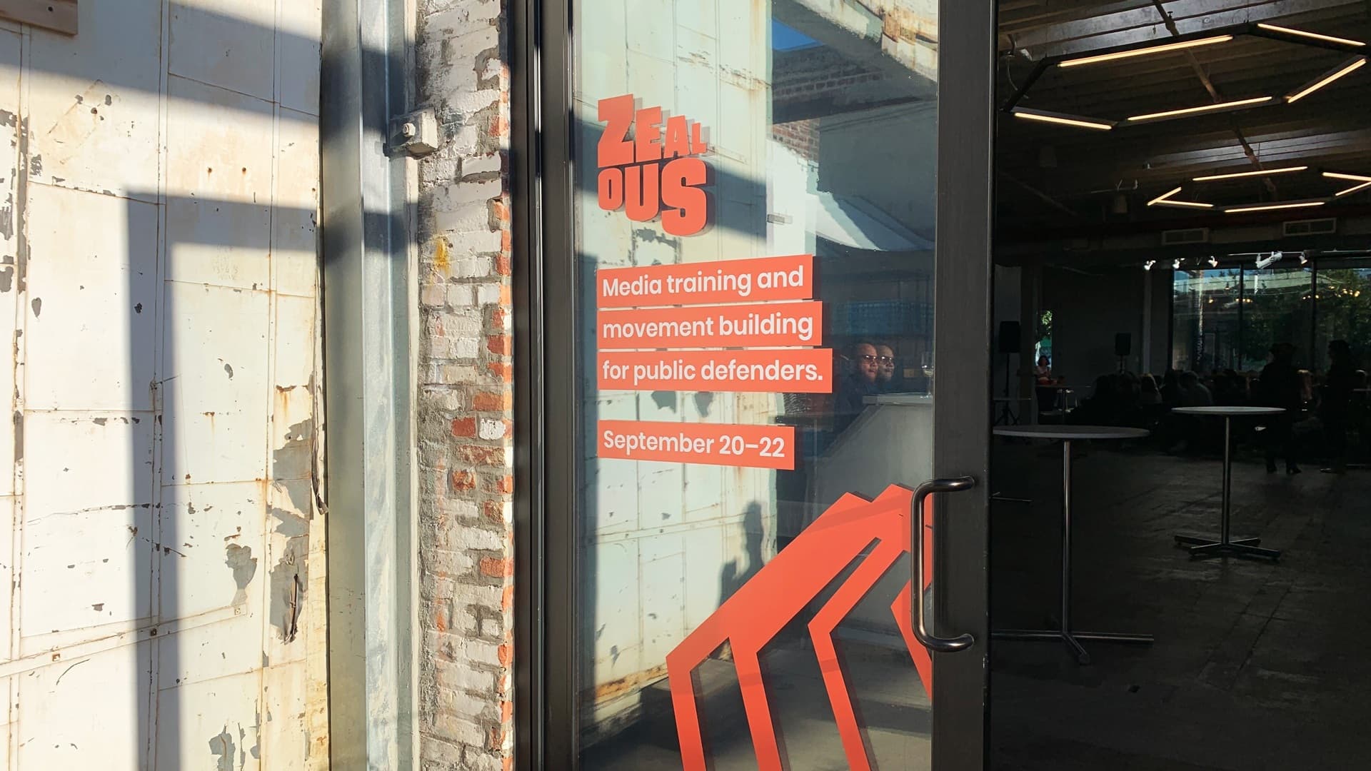 Glass door of a building with "Zealous: Media training and movement building for public defenders" in red and white text. Event dates "September 20-22" are also displayed. Partially visible interior shows tables and chairs under modern lighting.